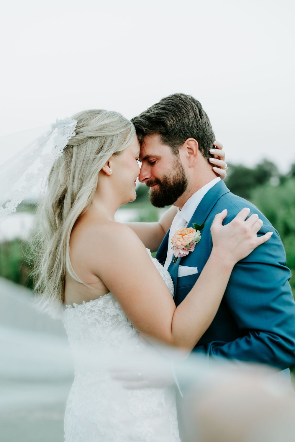 A bride and groom share a romantic forehead-to-forehead moment, with the bride in a lace gown and veil and groom in a blue suit.