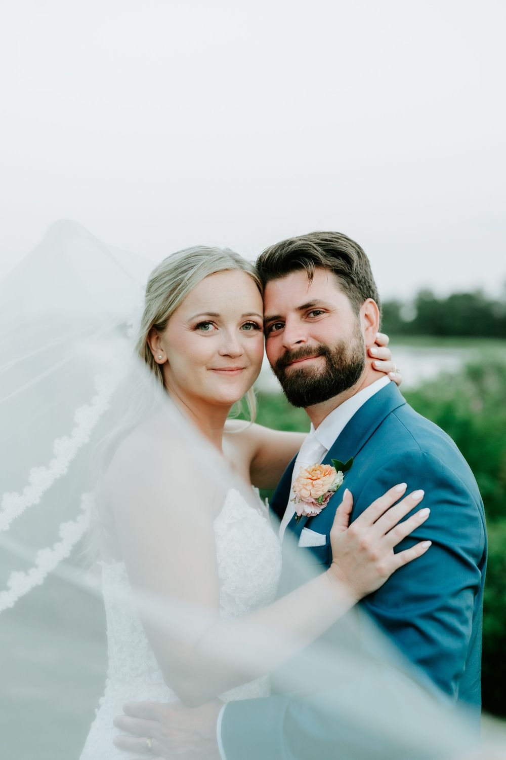 Bride and groom embrace under flowing veil in romantic outdoor portrait, groom wearing blue suit with boutonniere.