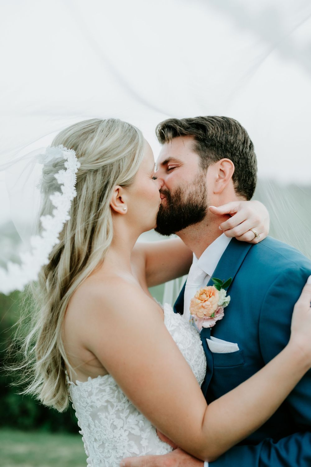 Bride and groom share a romantic first kiss, bride wearing lace gown with floral hair piece, groom in blue suit.