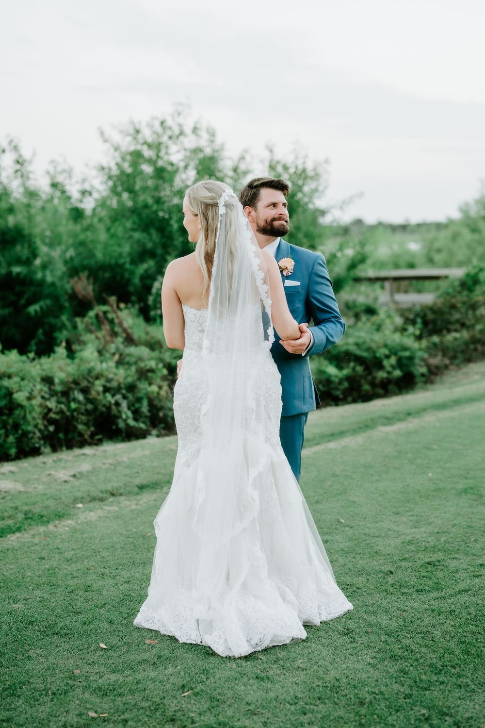 A bride in a white lace gown and veil stands with a bearded groom in a blue suit on a green lawn.
