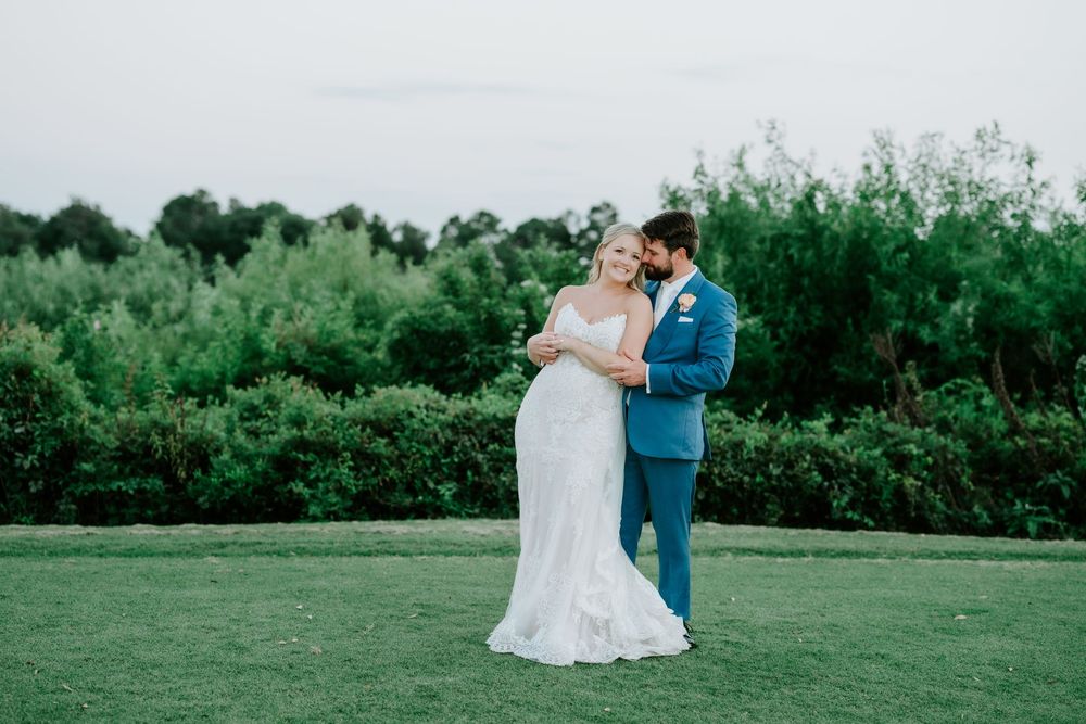 Bride and groom embrace on lush green lawn at outdoor wedding, surrounded by tall trees at dusk.