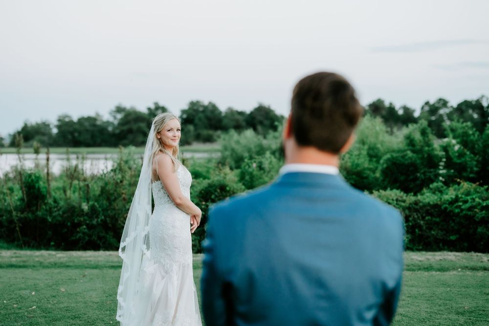 Bride in white lace gown and veil smiles over shoulder at groom in blue suit during outdoor wedding ceremony.