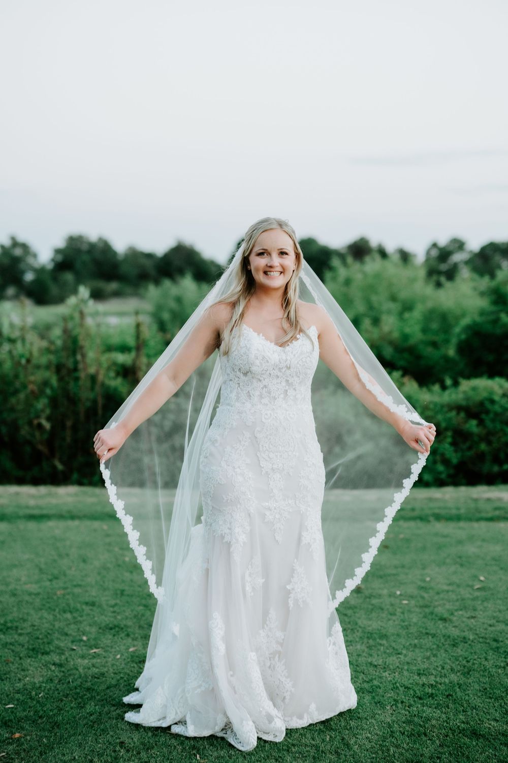Smiling bride in lace mermaid gown and cathedral veil posing outdoors on green lawn with trees in background.