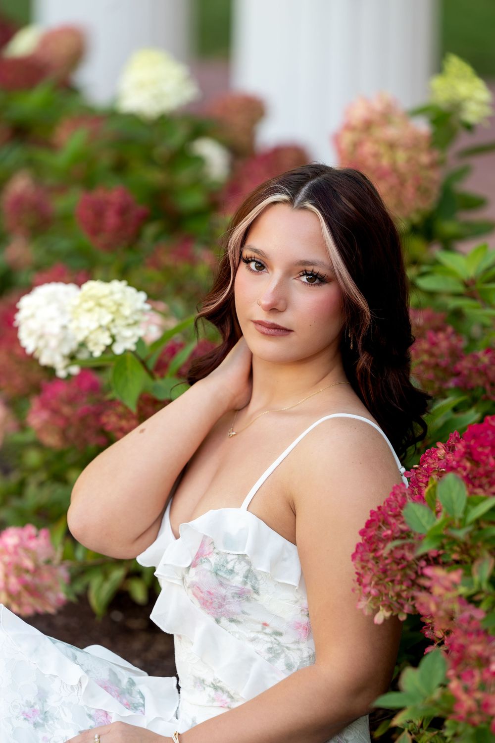 Young woman in white floral dress poses gracefully among pink and white hydrangea blooms in a lush garden setting.