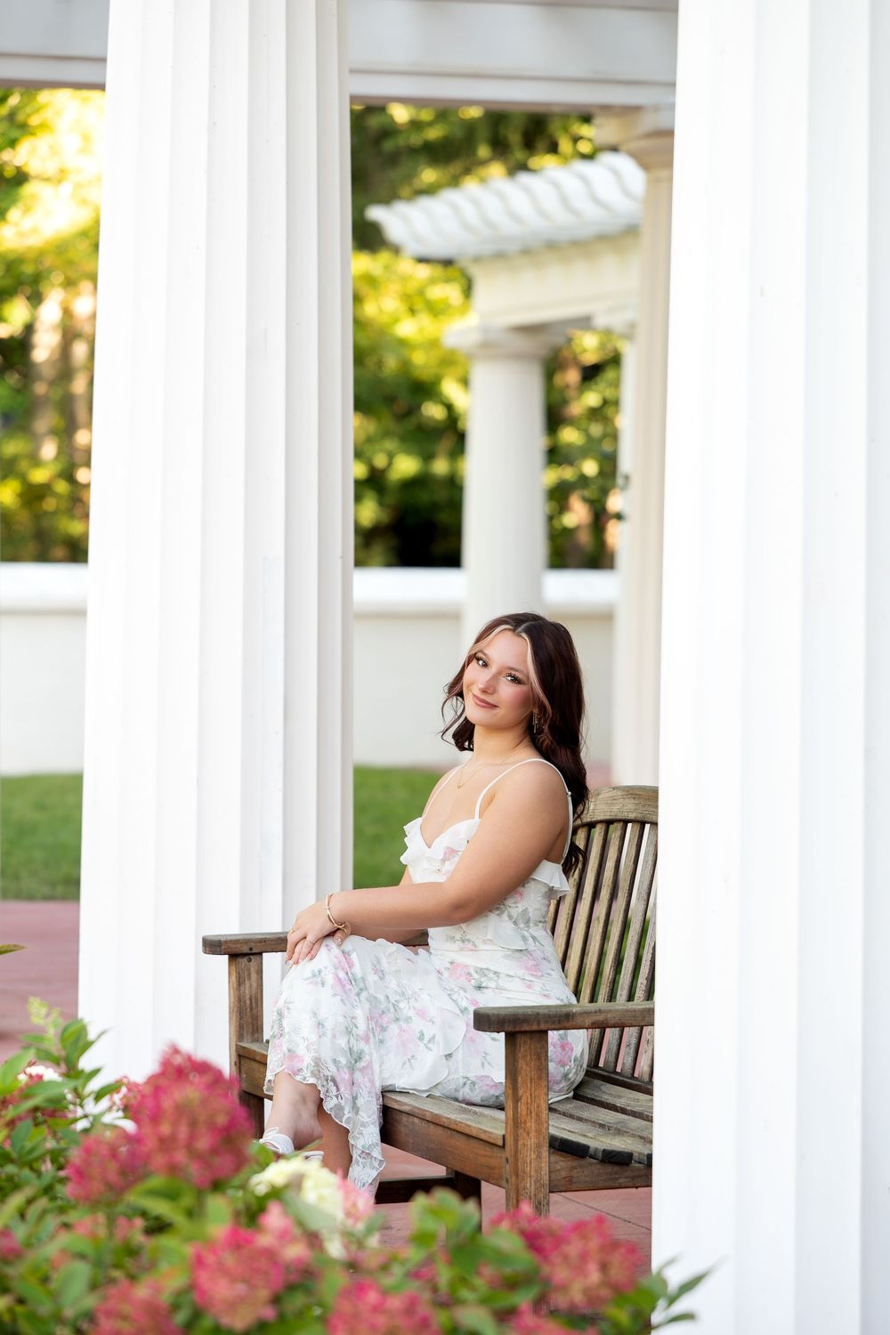 Brunette woman in floral dress seated on wooden bench between white columns in garden.