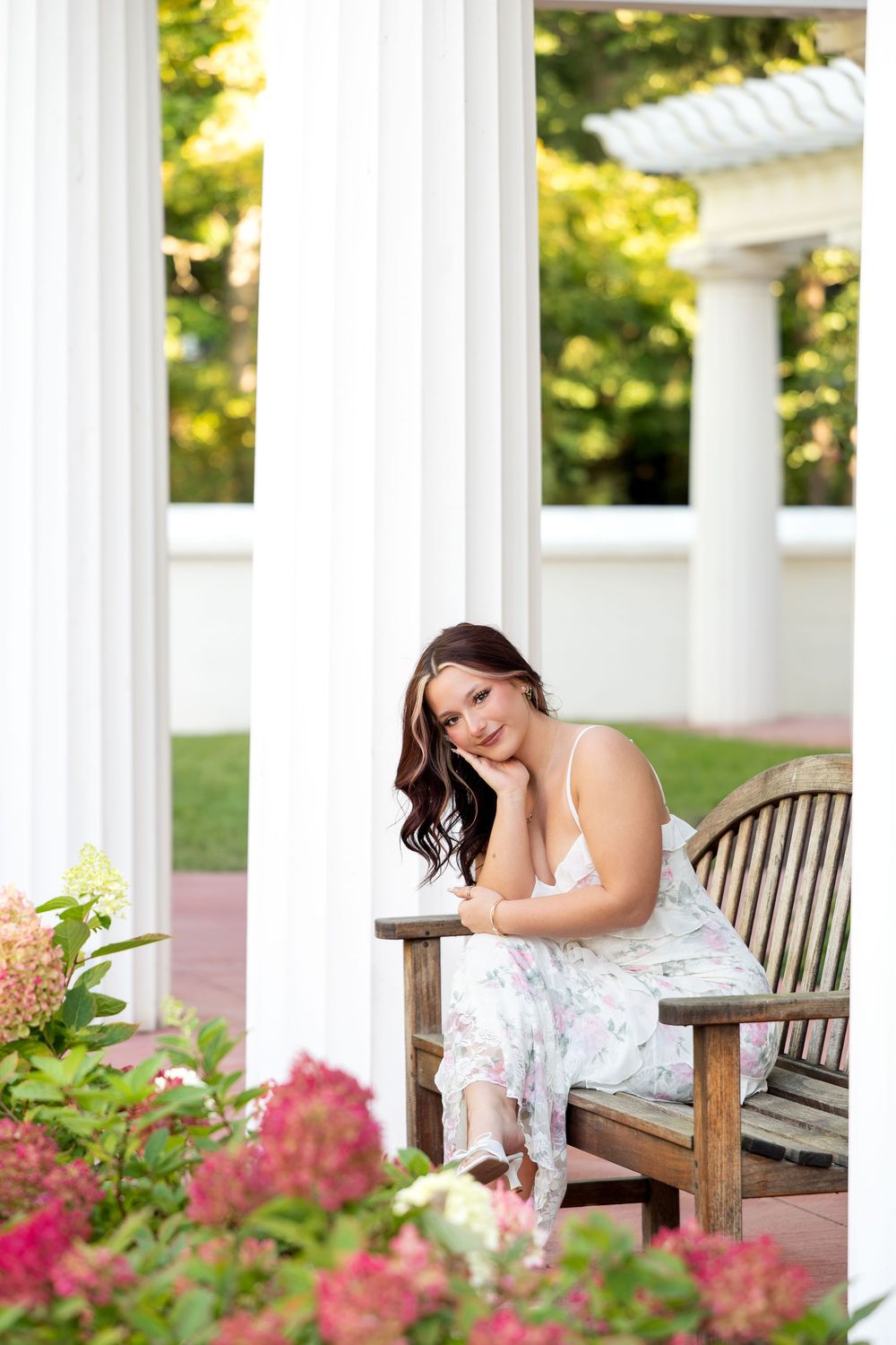Young woman in floral dress sitting on wooden bench among hydrangeas by white columns in garden.