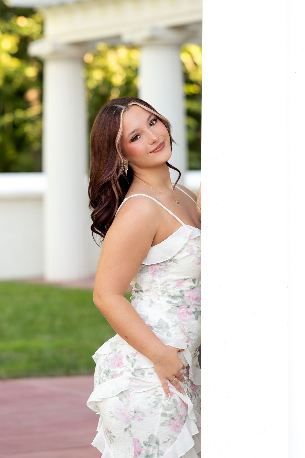 Young woman in floral spaghetti strap dress peeking around white column at elegant estate.