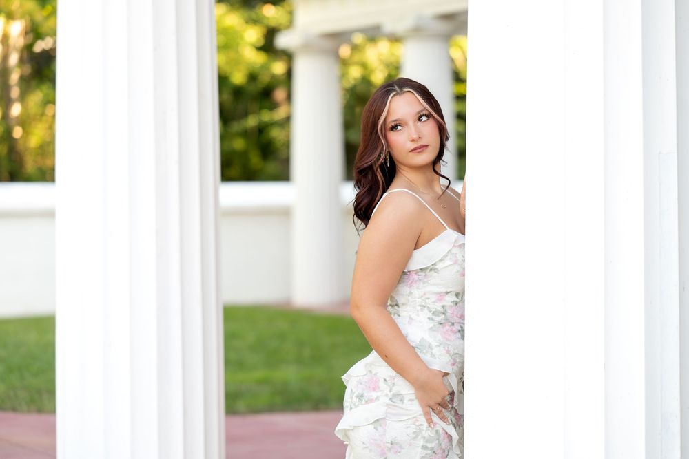 Young woman in floral dress standing between white columns looking up at senior portrait session.