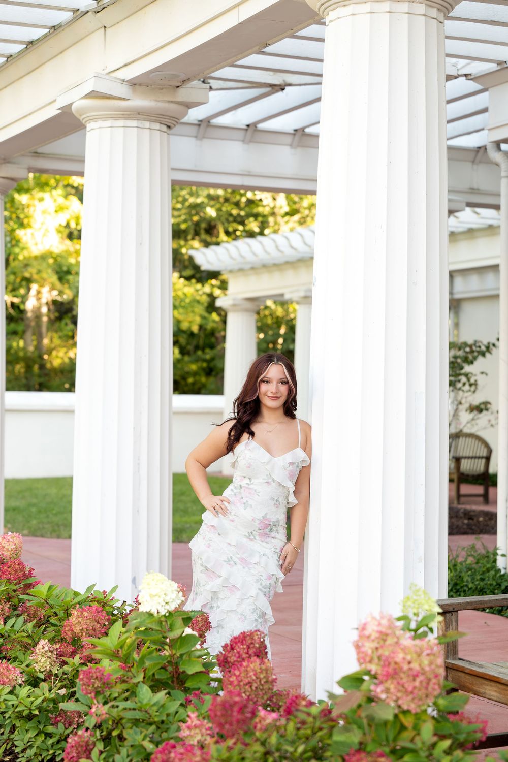 Young woman in white floral gown posing between tall white columns surrounded by hydrangeas.
