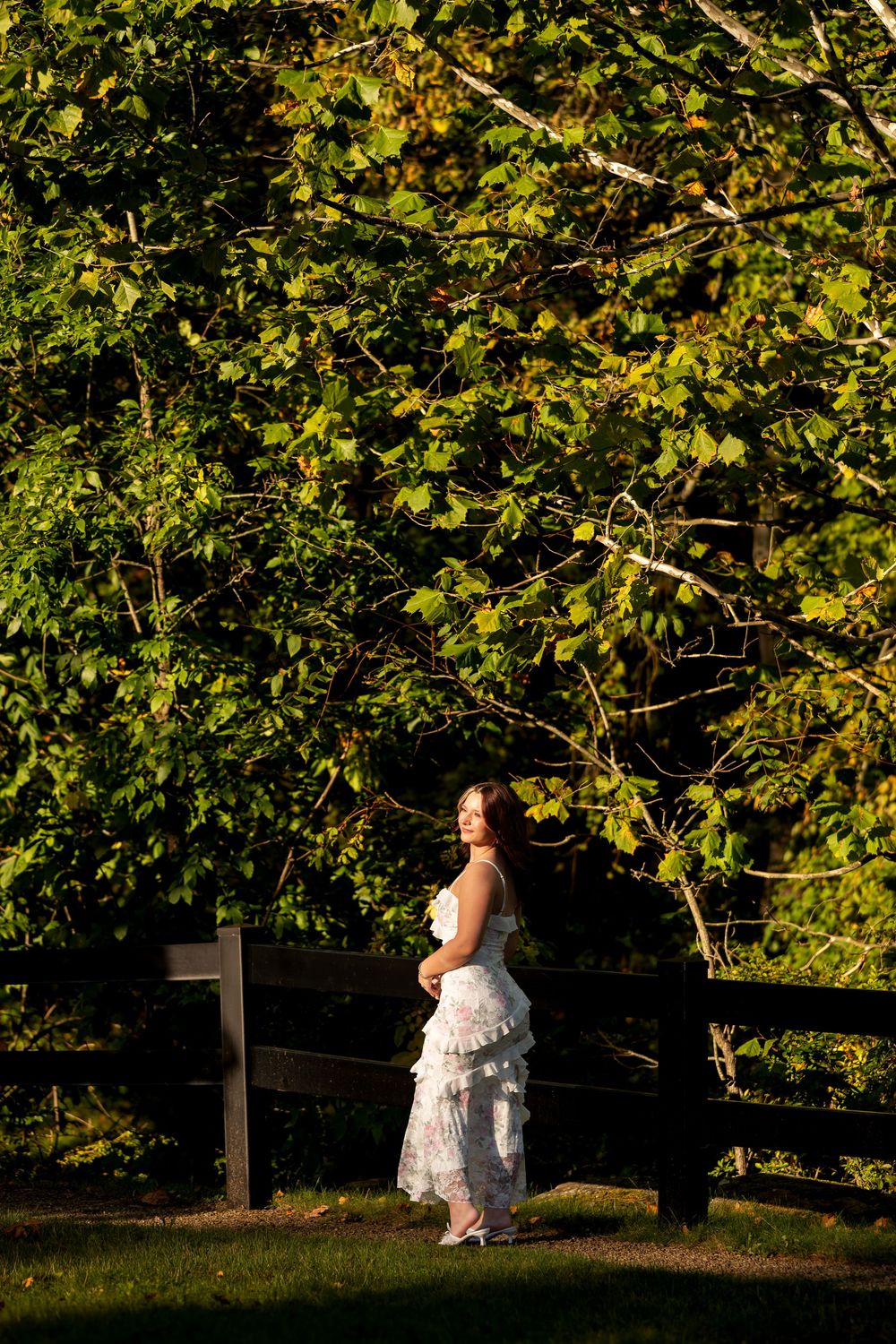 Elegant woman in white ruffled dress standing by wooden fence surrounded by green trees.