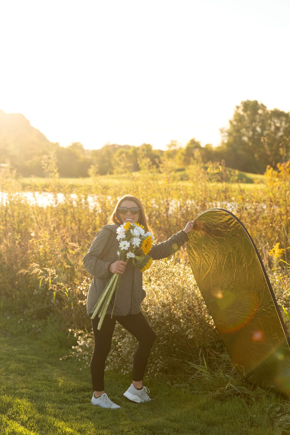 Woman in sunglasses holding white flower bouquet near reflector in sunny field.