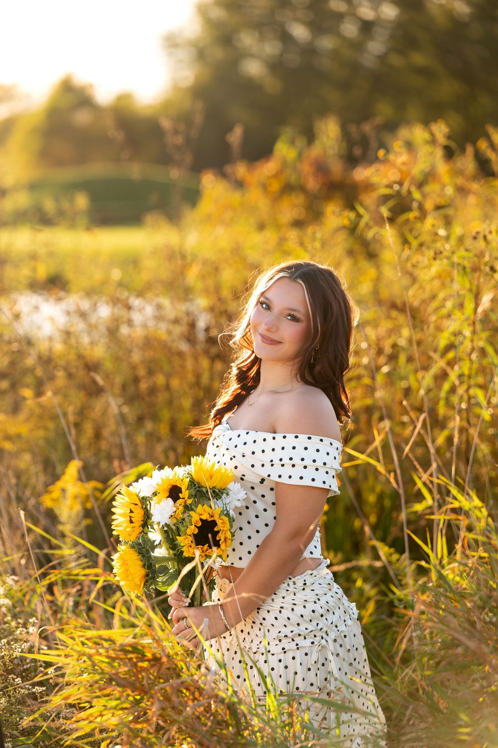 Young woman in polka dot dress holding sunflowers in a golden hour meadow setting.