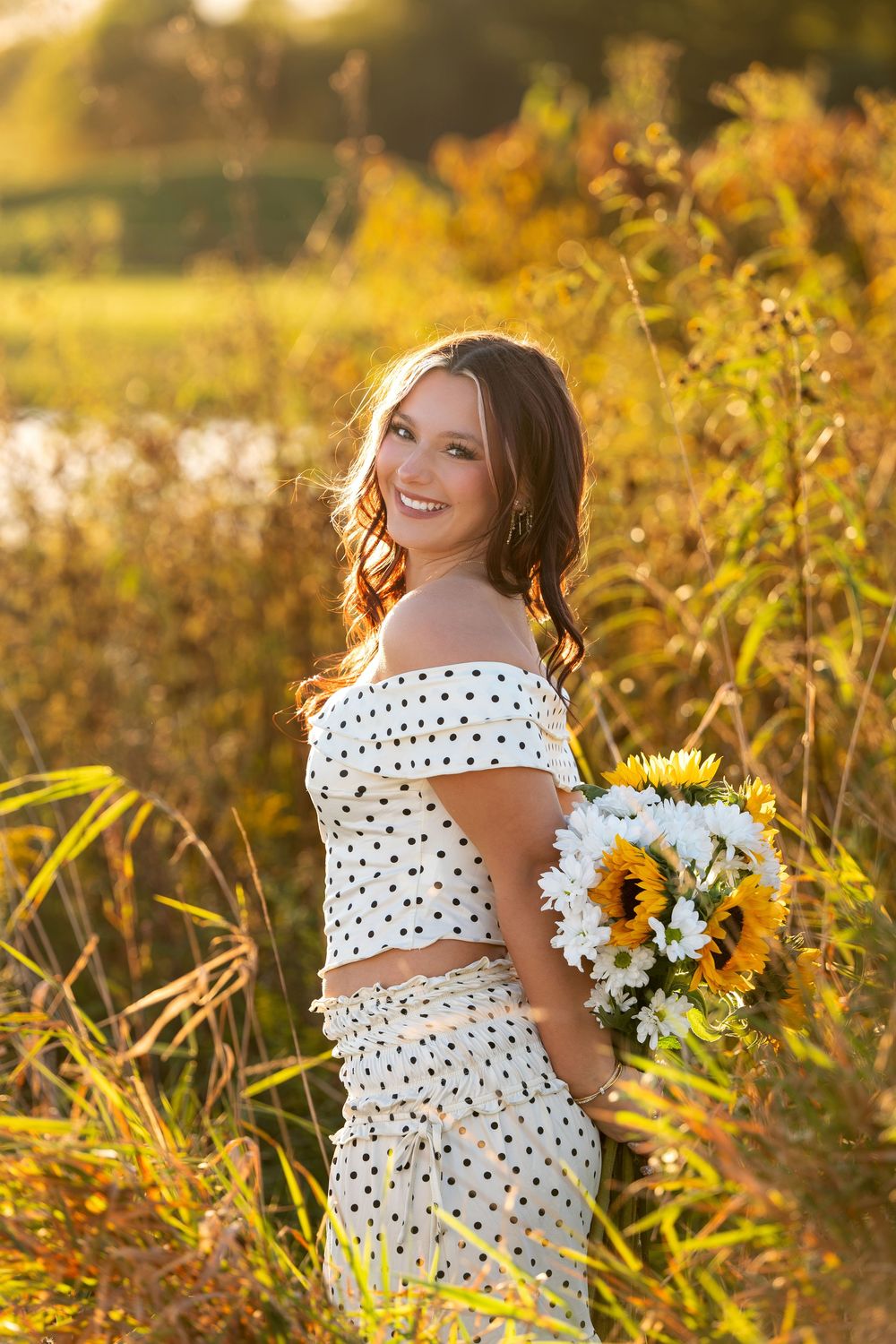 Joyful young woman in polka dot off-shoulder set holding sunflower bouquet in golden sunset field.