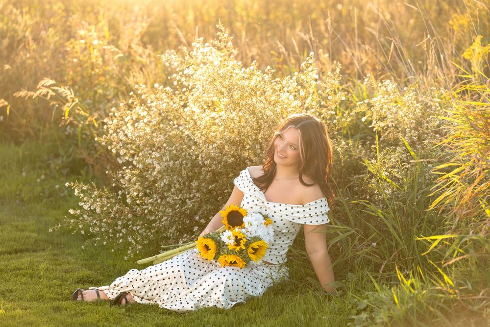 Young woman in polka dot dress sitting in golden field holding sunflowers at sunset.