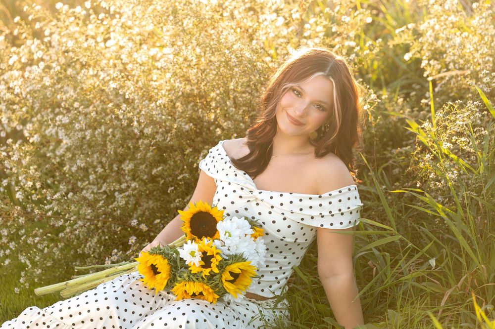 Smiling young woman in polka dot dress holds sunflowers while sitting in sunlit meadow.