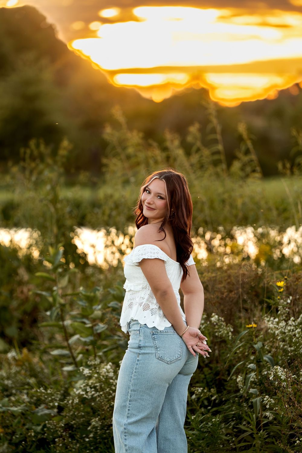 Young woman in white top and jeans posing in wildflower field with dramatic golden sunset sky.