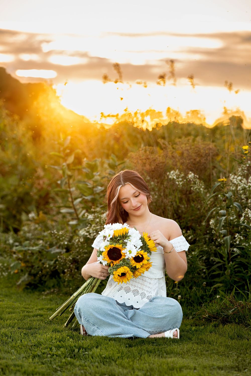 Young woman sitting cross-legged on grass holding sunflower bouquet at golden sunset.