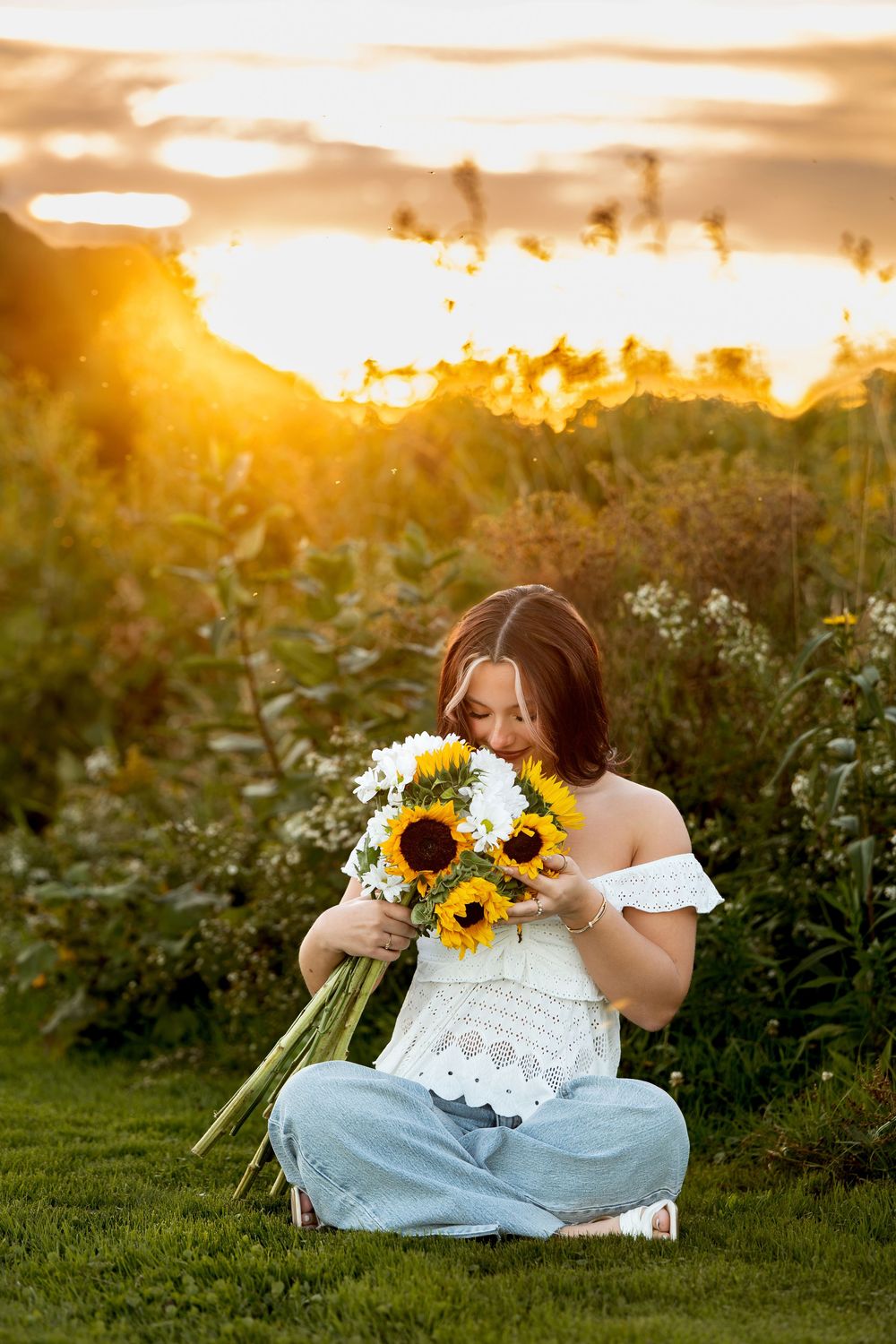 Woman smelling sunflower and daisy bouquet while sitting on grass at sunset.