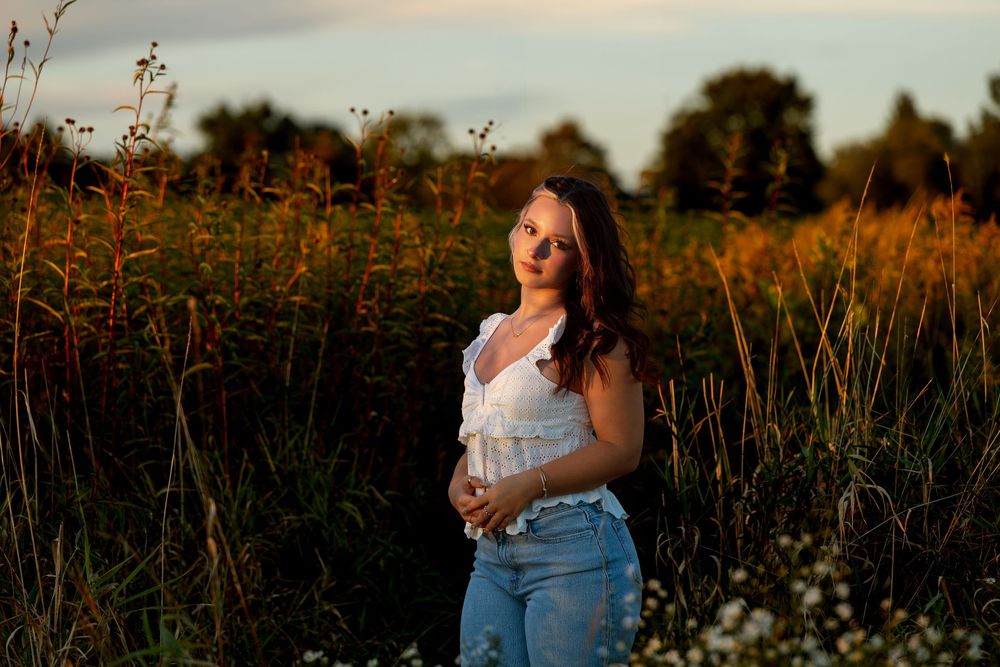 Pregnant woman in white top and jeans stands in golden field at sunset, cradling her baby bump.
