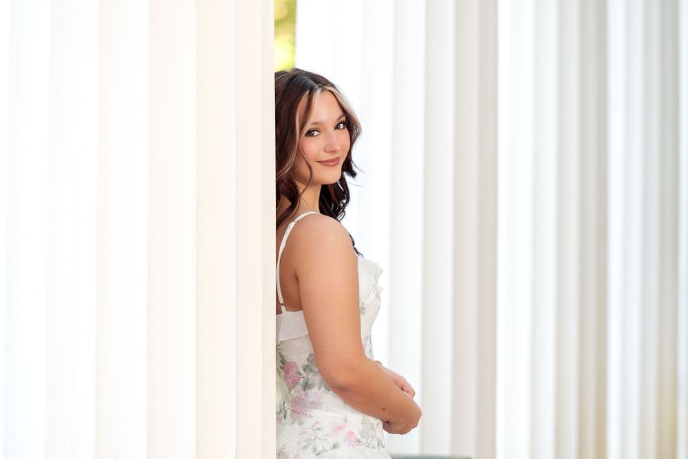 Elegant bride in a floral dress posing between white columns, smiling over her shoulder in a bright, airy setting.