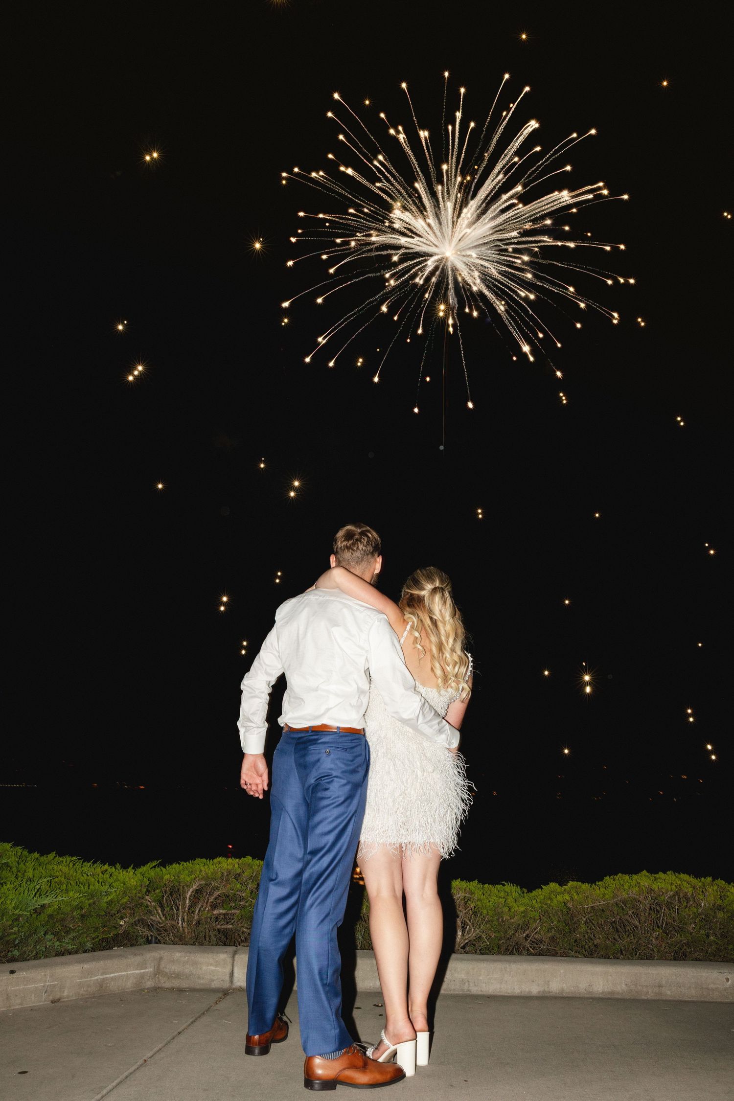 Bride and groom share a romantic kiss by a lakeside dock, groom in blue suit, bride in elegant white gown.
