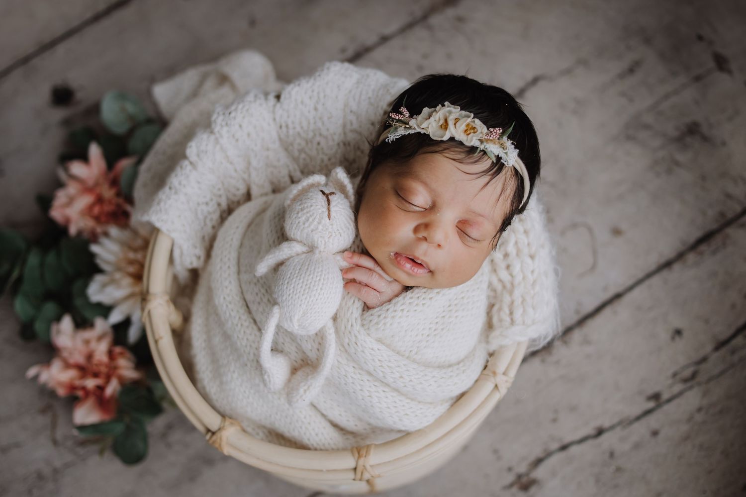 Sleeping newborn baby wrapped in white knit blanket, holding a bunny toy in a wooden bucket with floral headband.