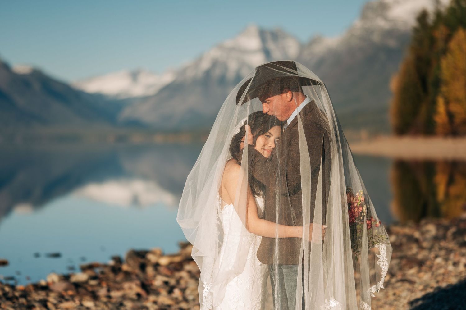 Bride and groom share romantic moment under veil by mountain lake with snow-capped peaks reflected in calm water.