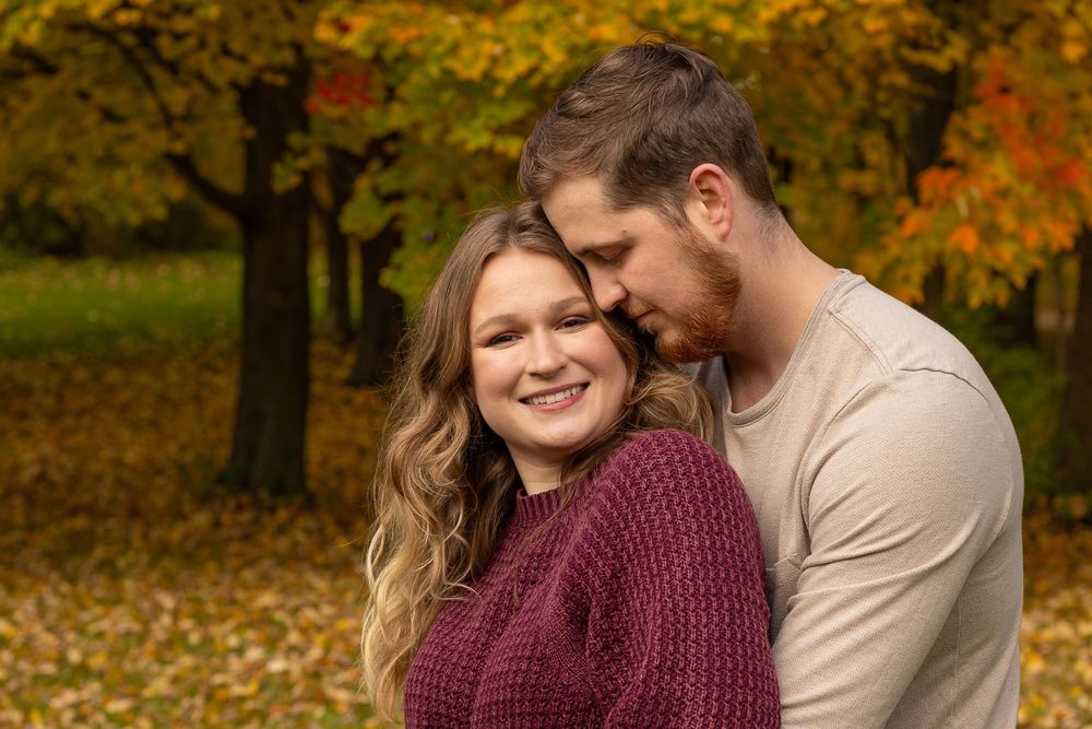 Couple embracing in autumn park, woman smiling in burgundy sweater, man kissing her temple, colorful fall foliage background.