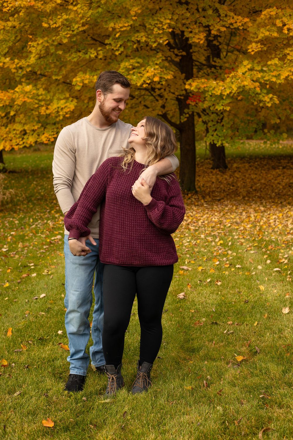 Couple embracing and smiling at each other in a park with vibrant autumn foliage and green grass.