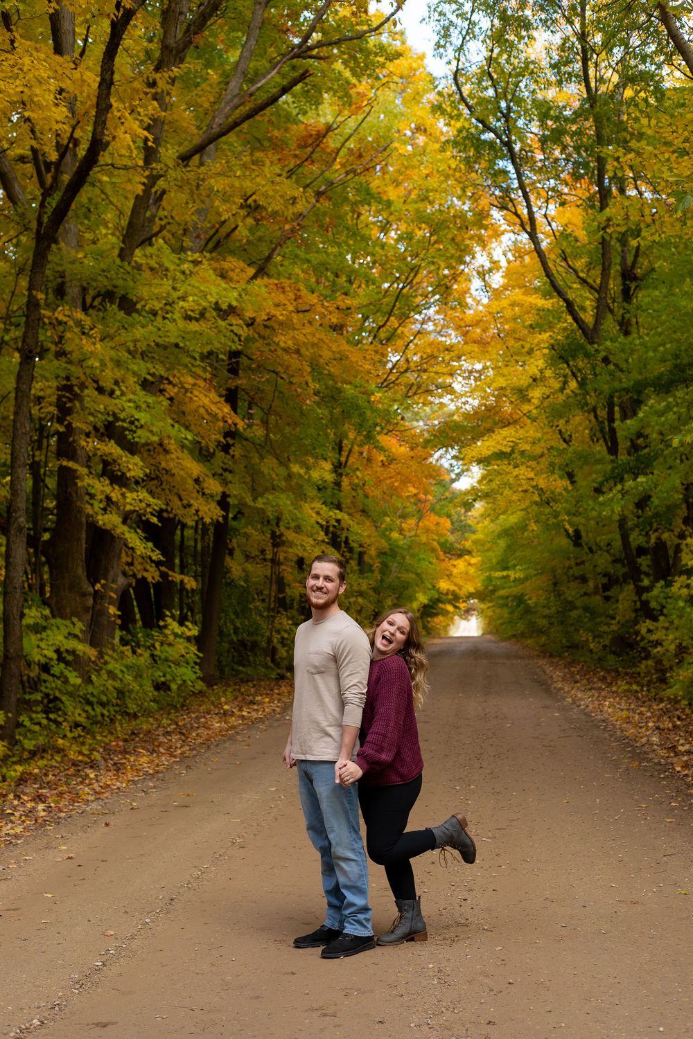 Couple smiling on a tree-lined dirt road surrounded by vibrant autumn foliage in yellow and green.