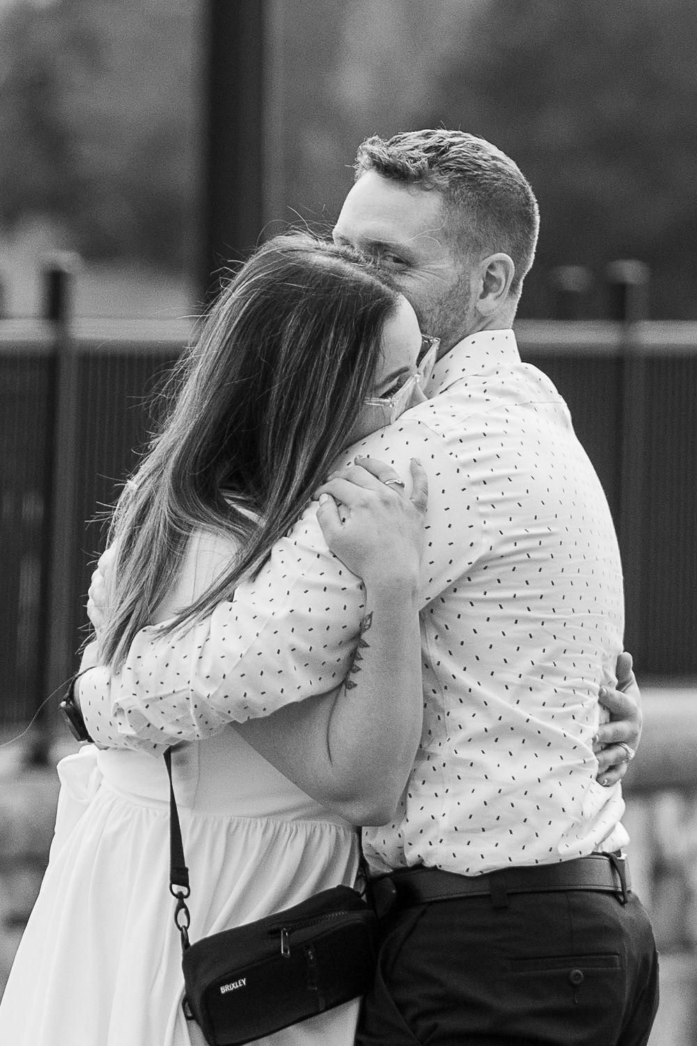 Black and white photo of a couple sharing a tender embrace outdoors, woman in polka dot dress hugging man in button-up shirt.