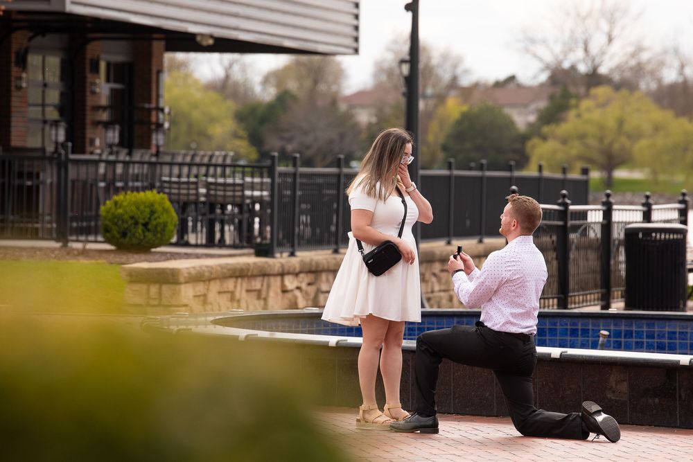 Man proposes on one knee to woman in white dress by outdoor fountain in a scenic park setting.