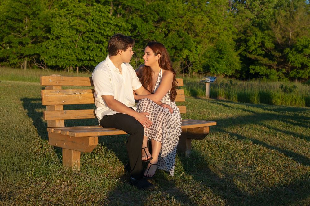 A couple sits closely on a wooden bench in a sunny park, gazing at each other romantically.