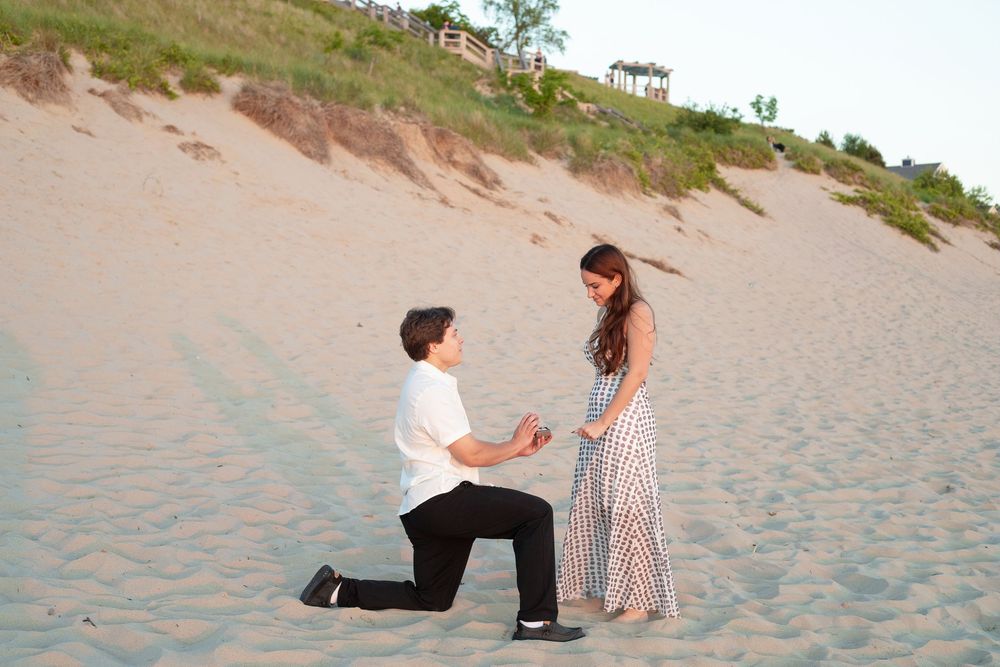 Man kneeling on sandy beach proposing to woman in striped dress at sunset near sand dunes.