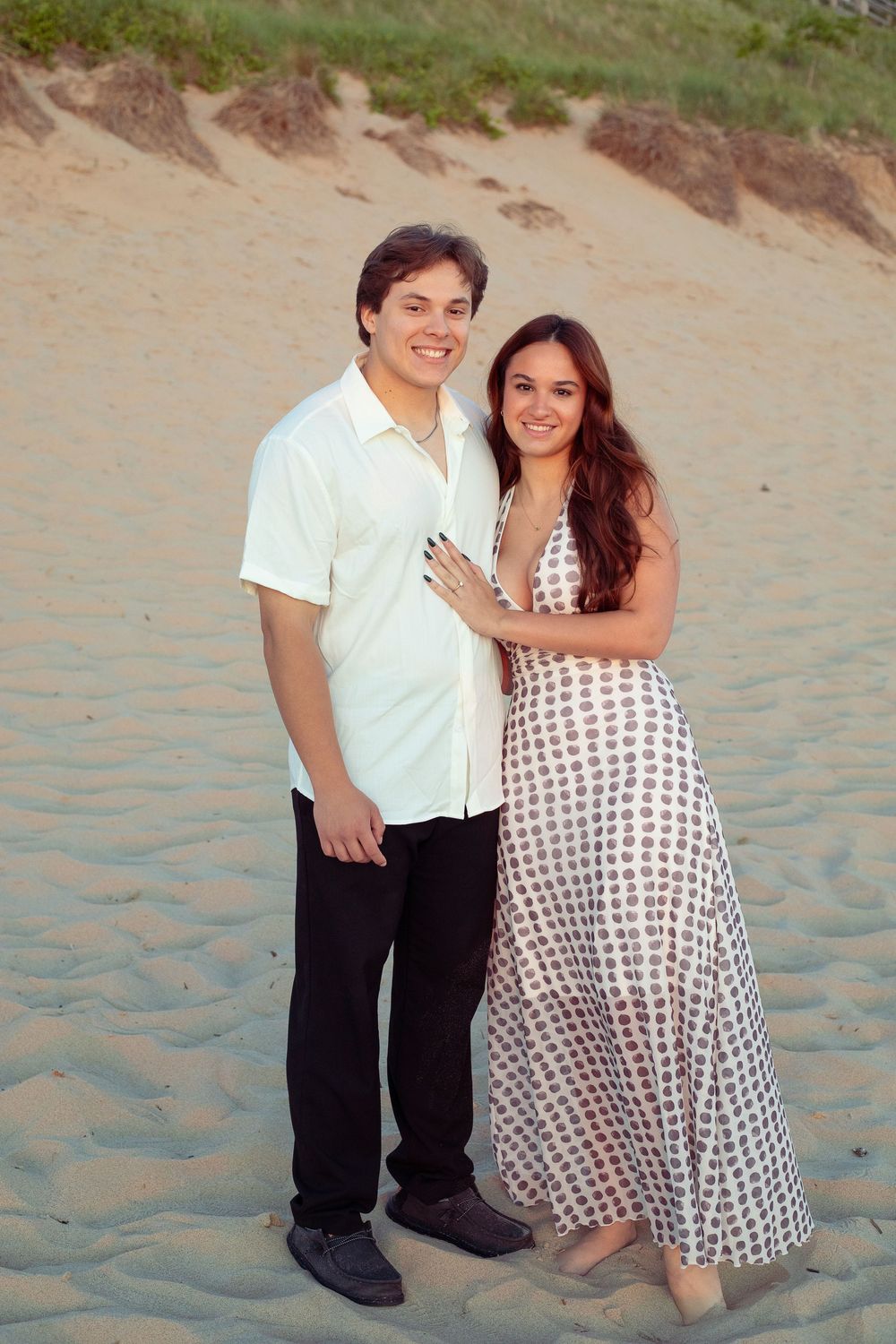 A smiling couple poses together on a sandy beach, the woman in a polka dot maxi dress with her hand on the man's chest.