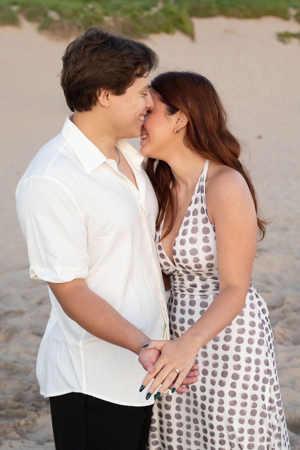 A couple shares a romantic moment on a sandy beach, the woman in a polka dot dress and man in a white shirt holding hands.