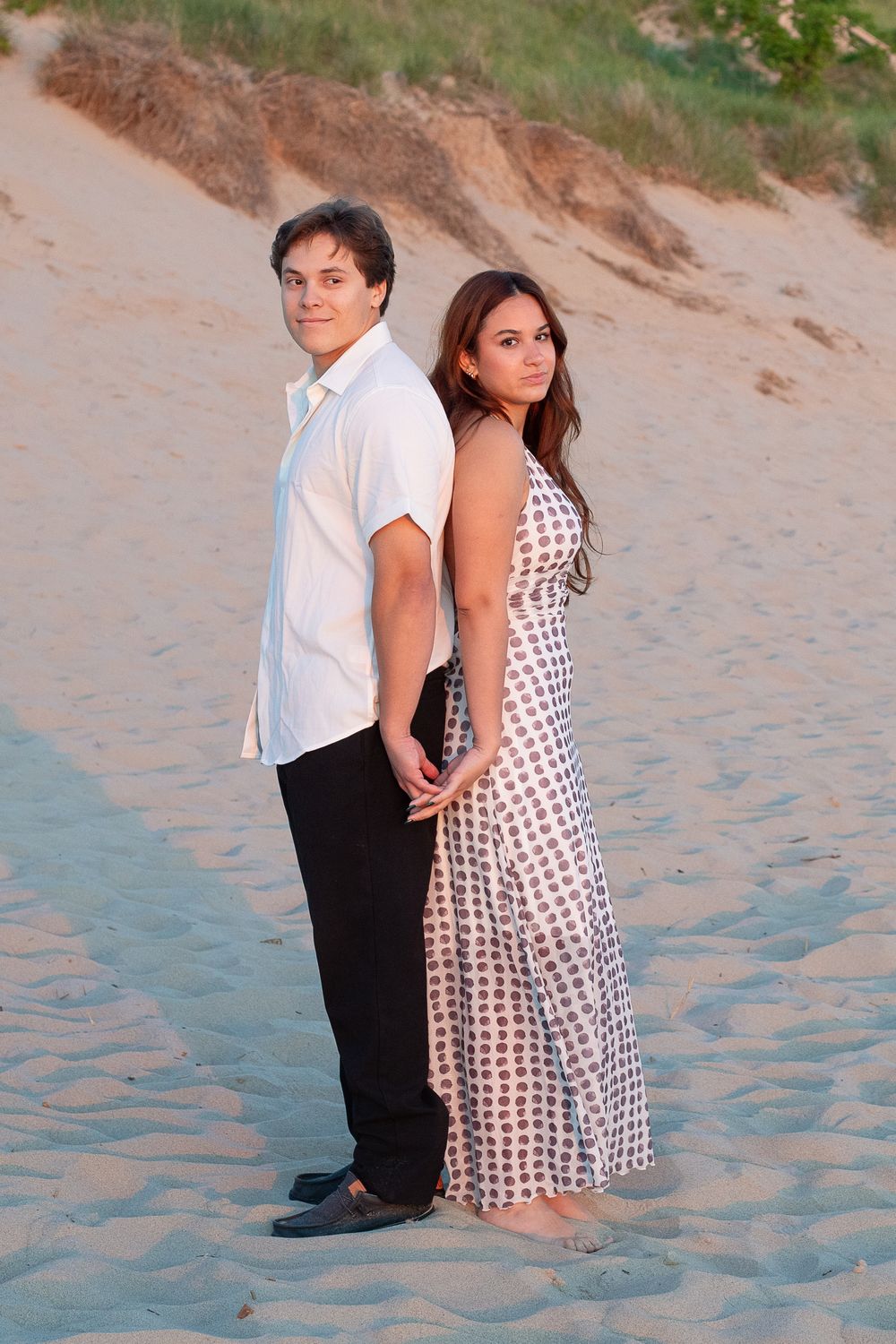 A young couple stands back-to-back on sandy beach, man in white shirt and black pants, woman in patterned maxi dress.