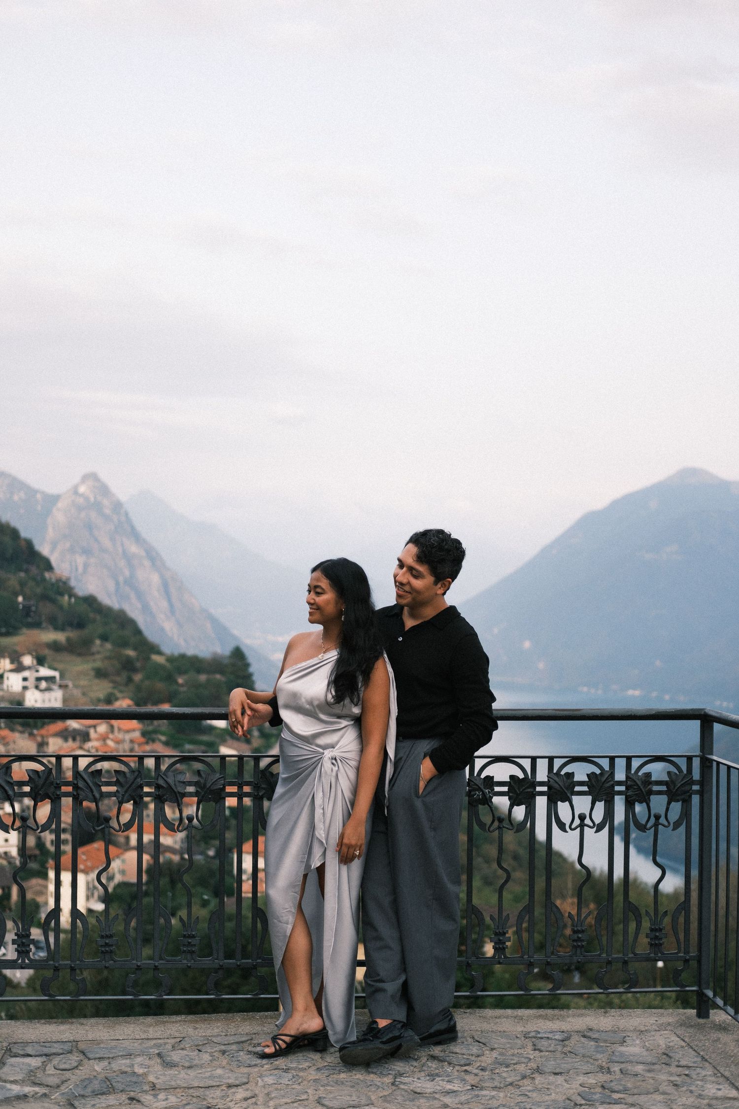Couple posing on a scenic balcony with mountain and lake views, woman in silver dress, man in black top.