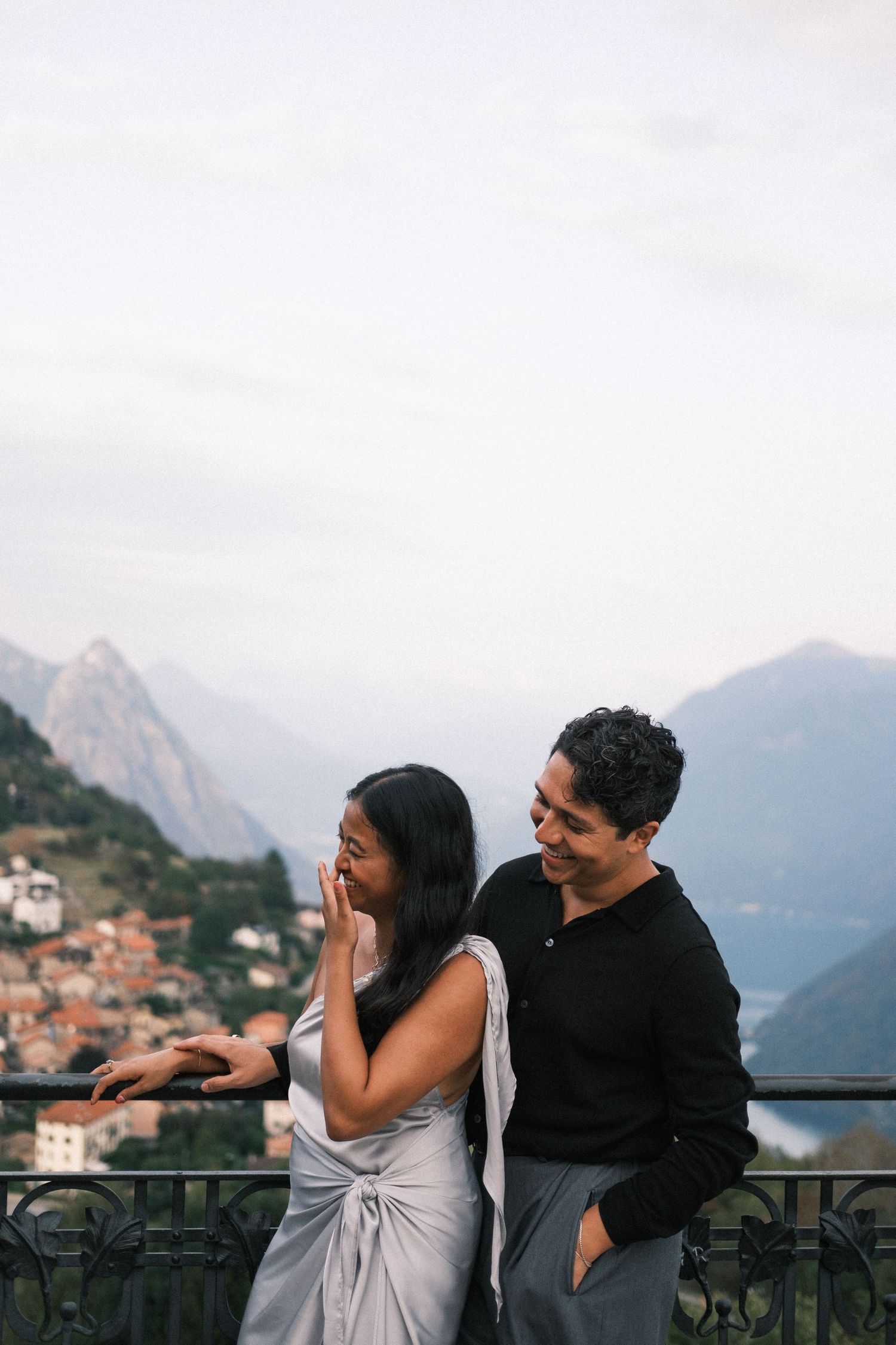 Couple laughing together on a scenic balcony with mountain and lake views in the background.