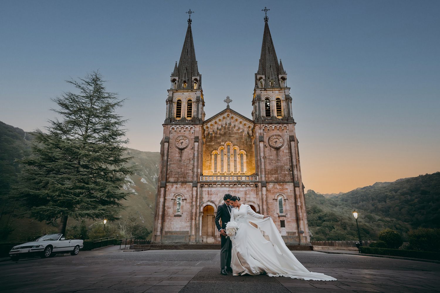 Novios frente a la fachada iluminada de la basílica de Covadonga al atardecer, con la cola del vestido extendida.