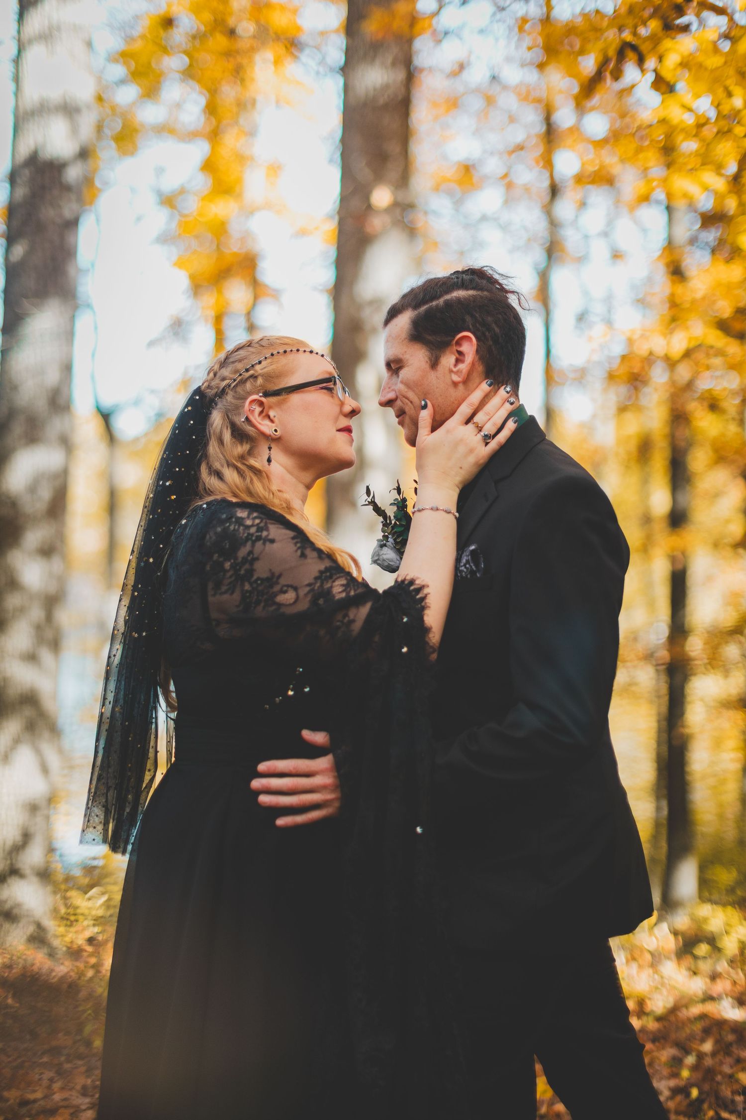 Couple in matching black wedding attire sharing a romantic moment in an autumn forest with golden foliage.