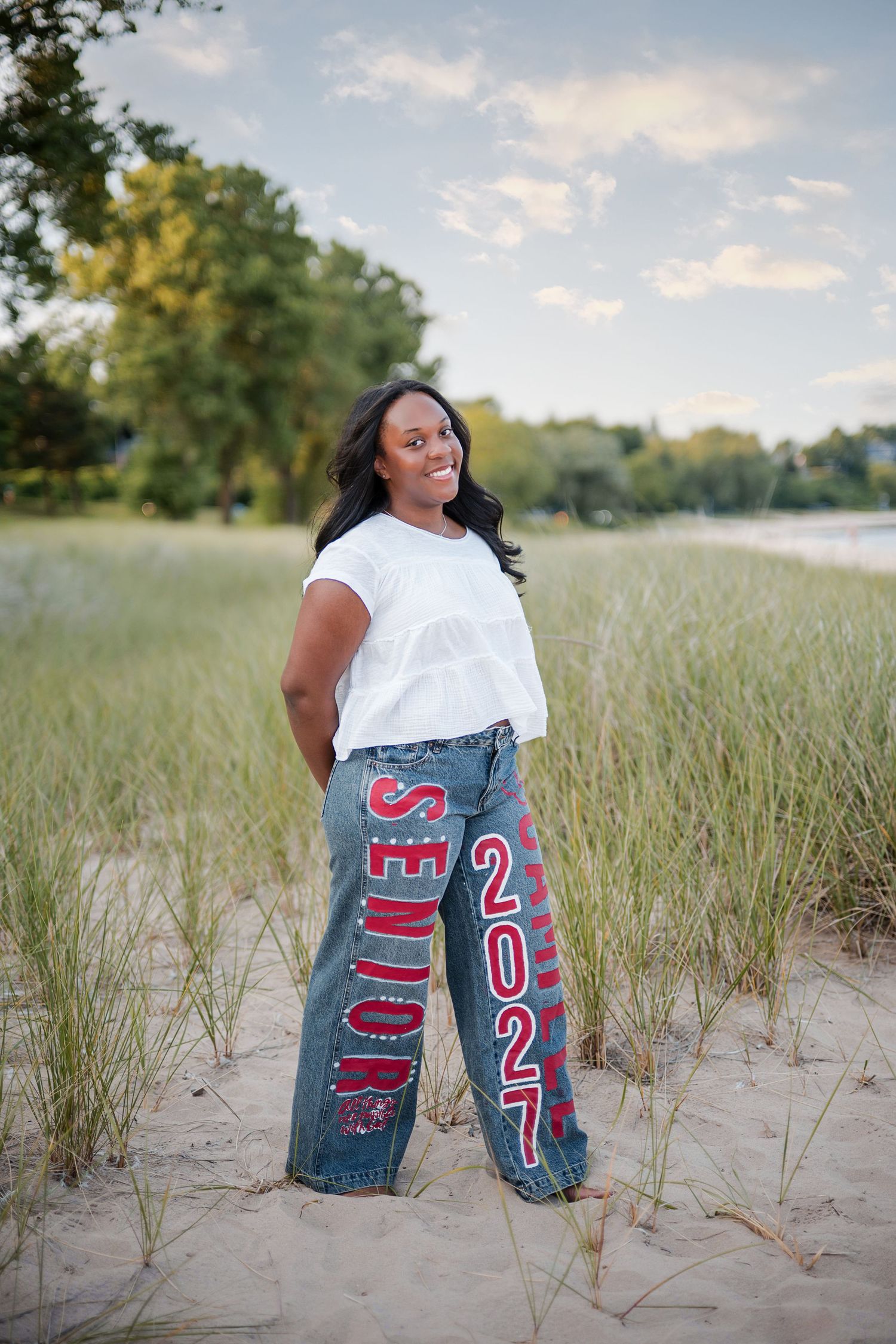 Smiling senior student poses on sandy beach dunes wearing custom painted 'Senior 2027' jeans and white top.