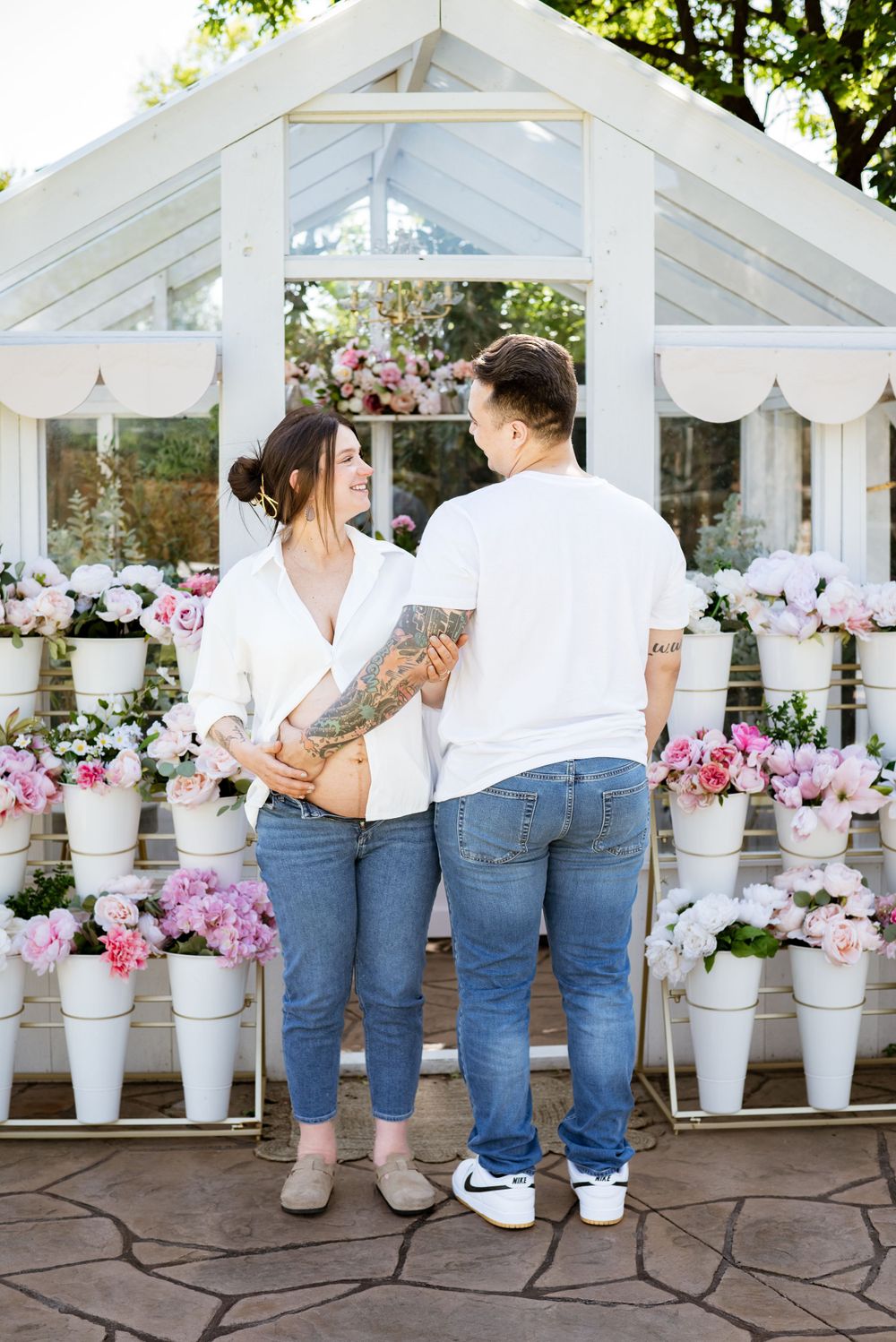 Couple in matching white tops and jeans posing by pink flowers in a greenhouse.