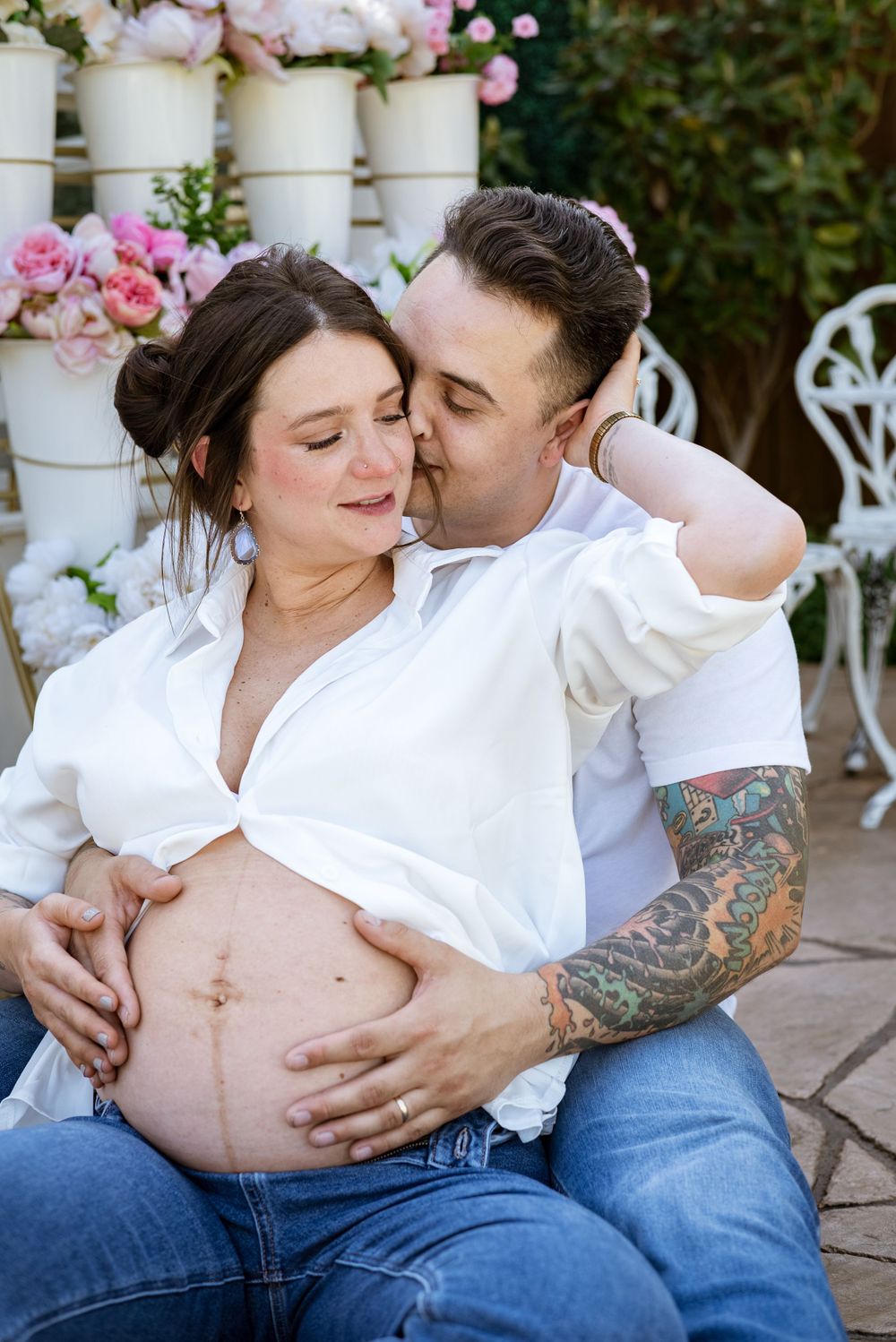 Intimate maternity photos of couple in white shirts and jeans surrounded by pink flowers.