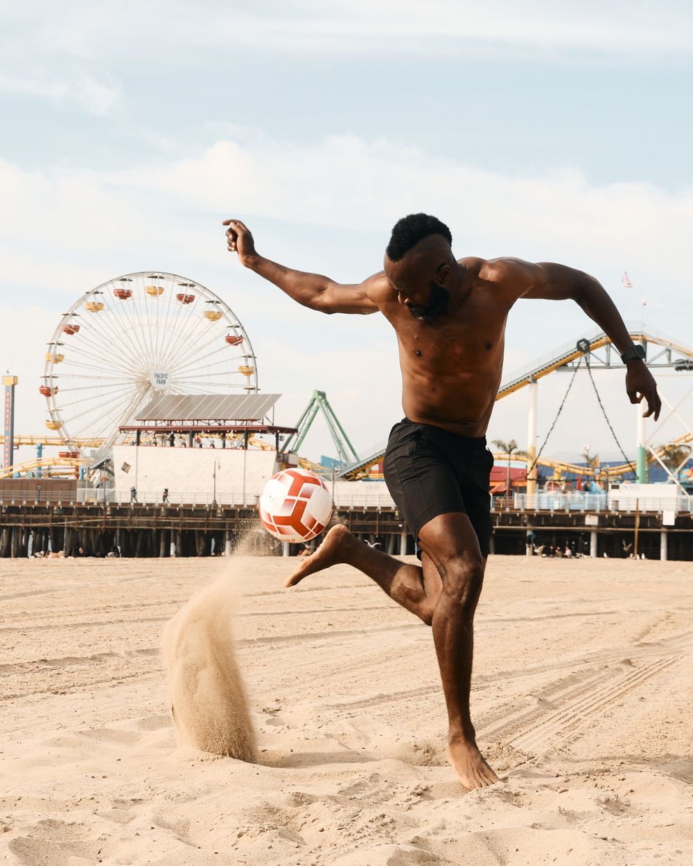 Athletic figure flicking up a soccer ball and kicking up sand on a beach with a ferris wheel and amusement park pier in the background.