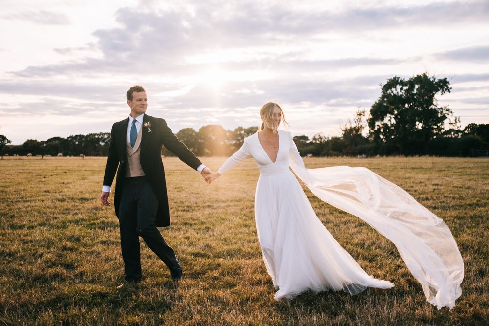 Wedding couple runs through a grassy field at sunset with bride's veil flowing in the wind.