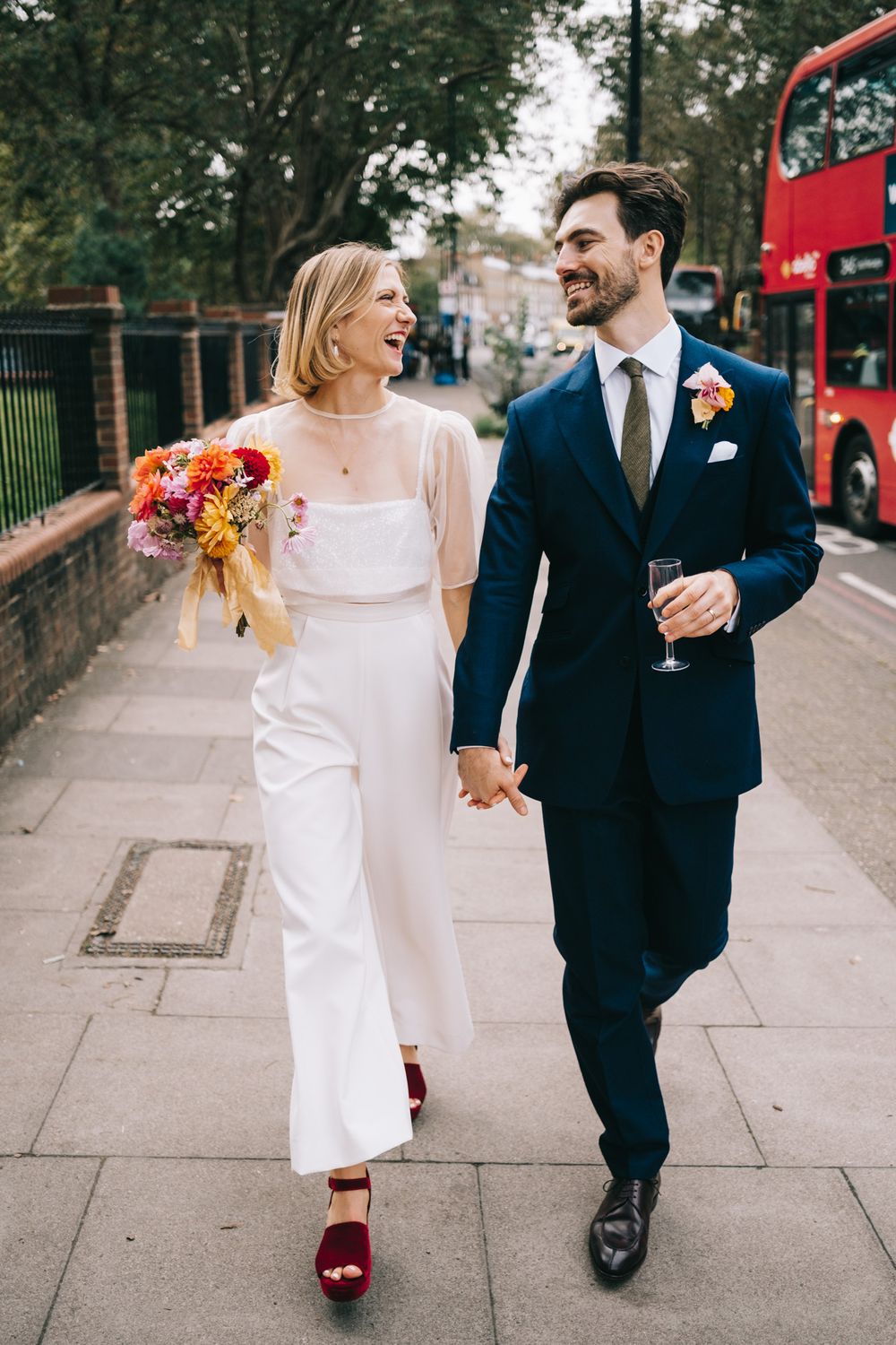 Newlyweds walk down a London street holding hands, bride carrying colorful bouquet.