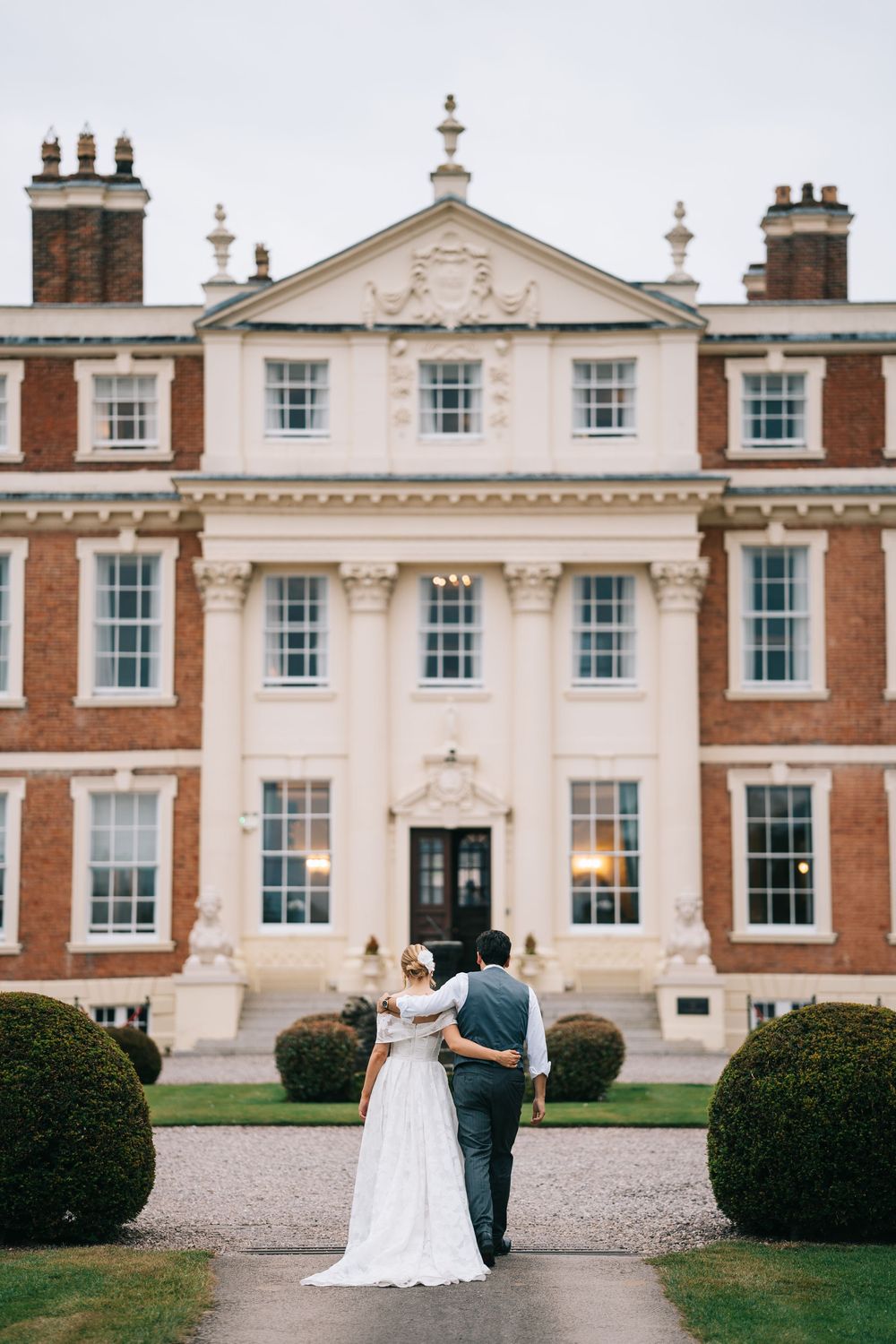 Elegant wedding couple in front of historic mansion with white columns and red brick exterior, Hawkstone Hall, Shropshire