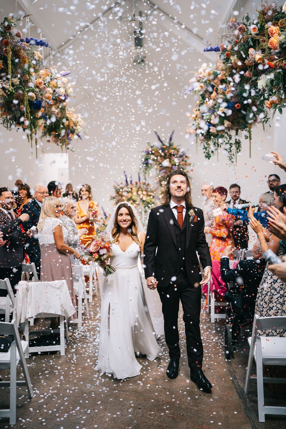 Newlyweds walk down the aisle under beautiful floral arrangements as guests shower them with confetti at fivefourstudios, Salford