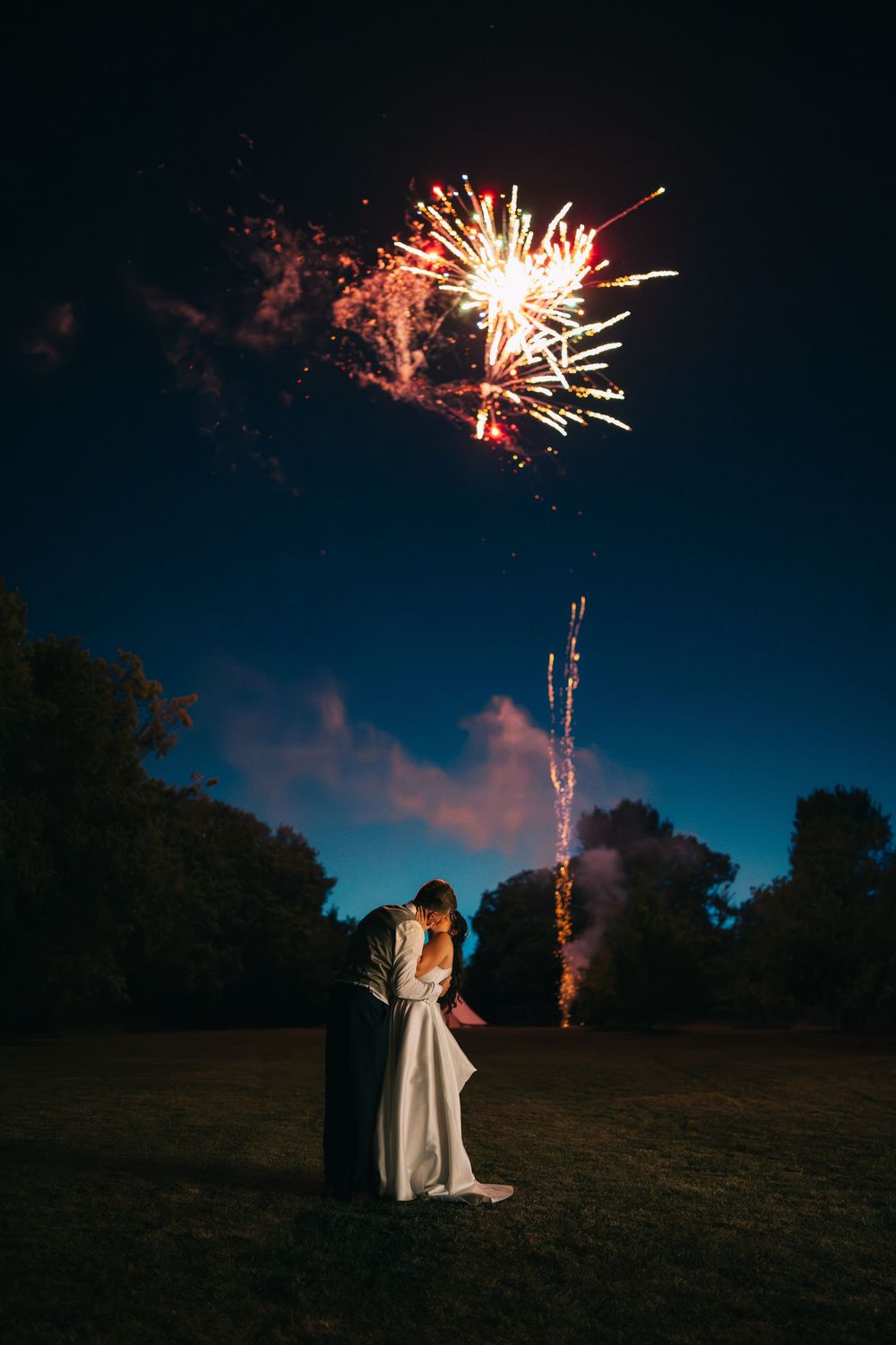 Silhouetted couple stands in a field at dusk as colorful fireworks burst in the dark blue sky above them.